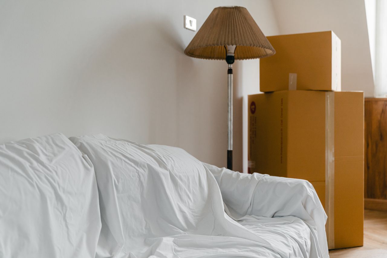 heros-img Covered with sheet sofa and floor lamp and packed cardboard boxes in light spacious room of new contemporary house