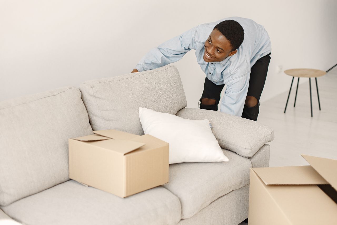 about-us-02 Young man smiling while arranging cardboard boxes in a modern living room.